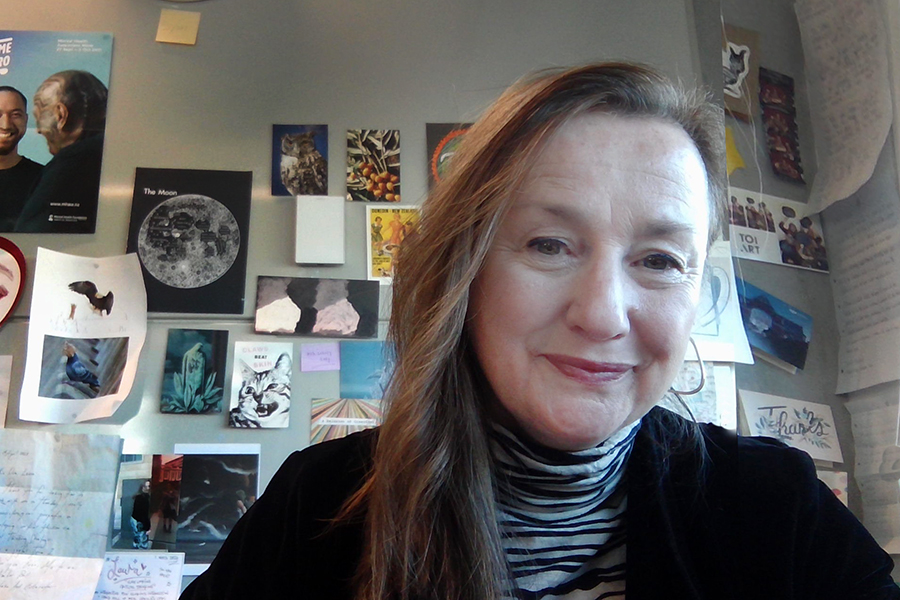 A close headshot of a woman with long hair drawn to one side. She is smiling at the camera. There are notes and postcards pinned to the wall behind her.