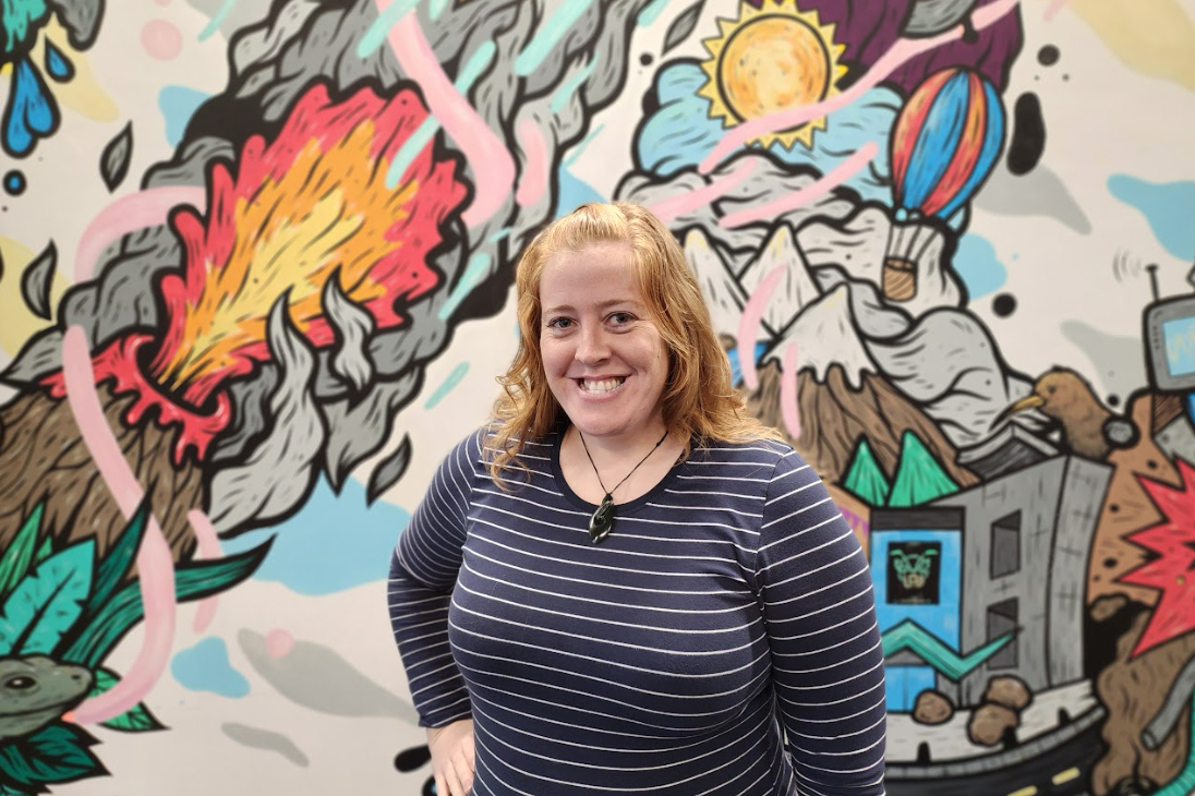 A woman in a blue and white striped top with long hair is smiling at the camera. She is standing in front of a colourful mural.