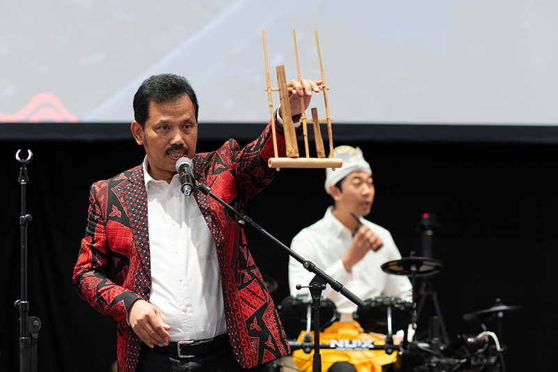 A performer stands on stage with a microphone stand holding and bamboo angklung. A percussionist stands behind the performer