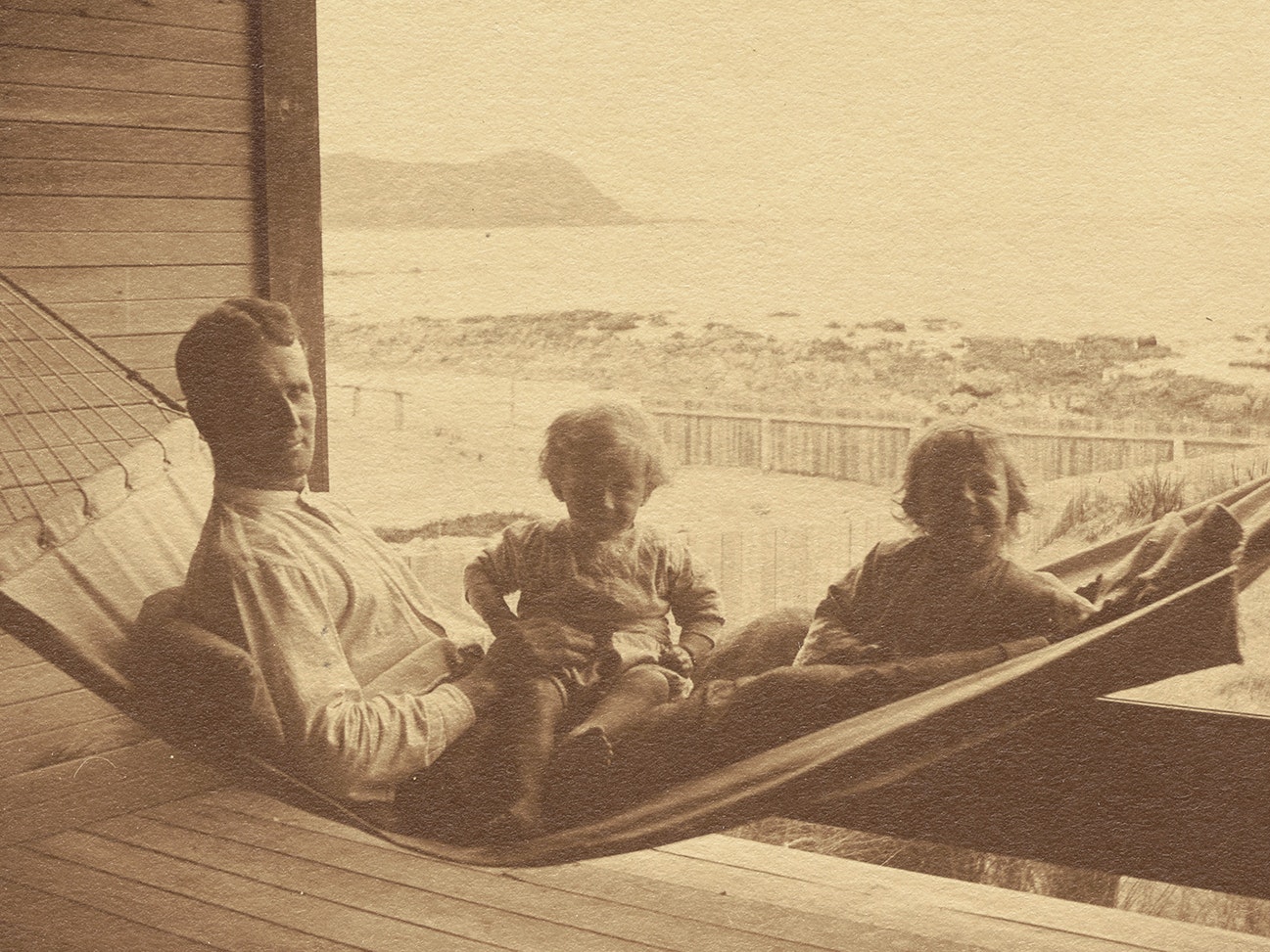 "Here we are again": 'Visit to Pukerua Bay, Feb. 11. 1920'. From the album: Family photograph album; 1917 - 1920; Adkin, Leslie, 11 February 1920, New Zealand, by Leslie Adkin. Gift of Adkin Family, 1997. Te Papa (O.031666) A sepia photo of a man in a hammock with two small children on his lap.