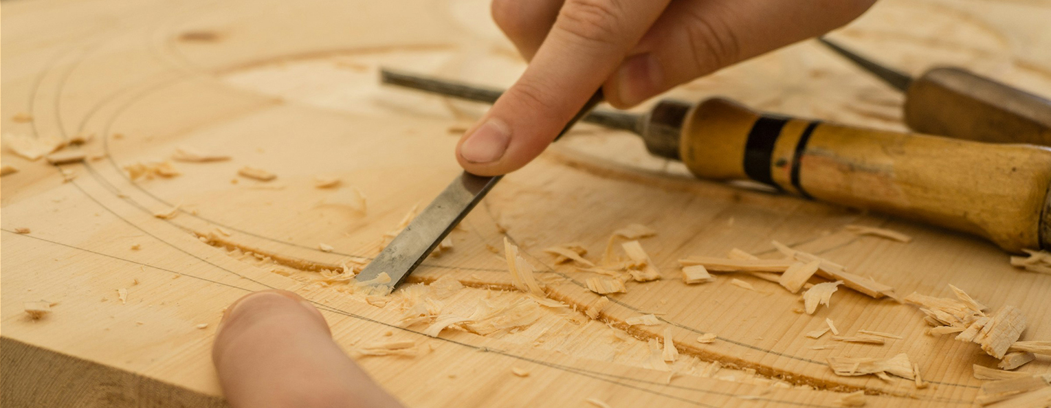 A closeup photo of two hands. One is holding a carving instrument ans is carving a piece of wood.