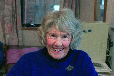 An older woman is sitting at a desk with plant illustrations in front of her. She is smiling at the camera.