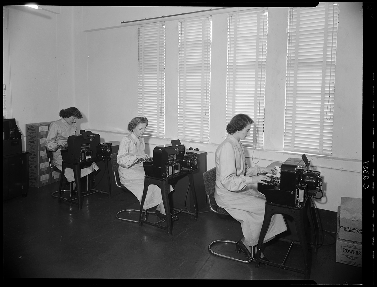 Black and white photo of Three unknown women using office equipment. Boxes of accounting machine cards are piled up on the floor.