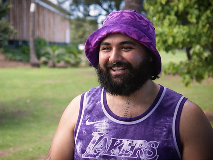 Still from Doron Semu video for ‘Ahu: Ngā Wairua o Hina A man in a purple bucket hat and a purple singlet is talking to someone off camera.