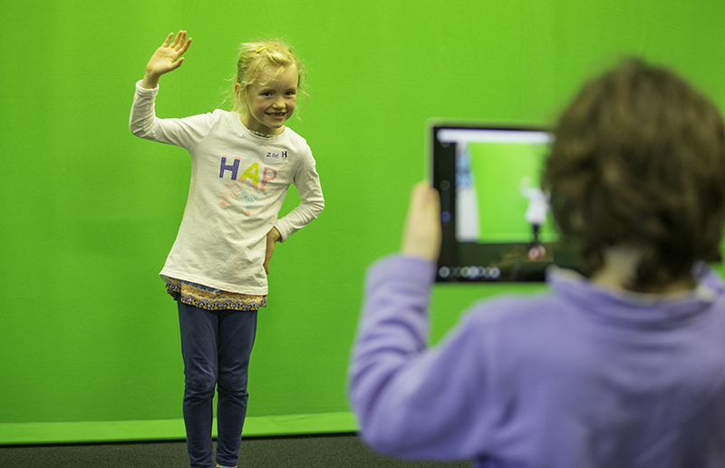 One child is standing in front of a bright greenscreen and waving and another is holding a tablet up to film her.