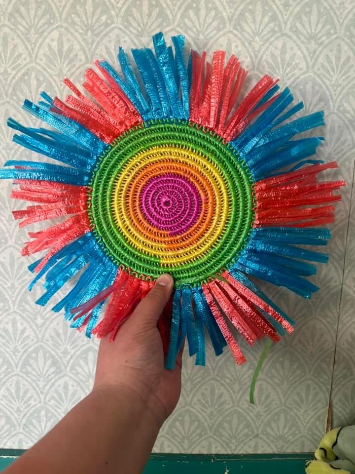 A hand is holding a colourful woven mat with pink and blue rafia fringe