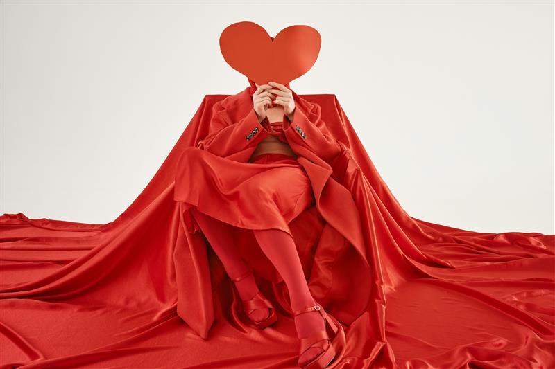 A person in red holding a red heart in front of their face is surrounded by red material.