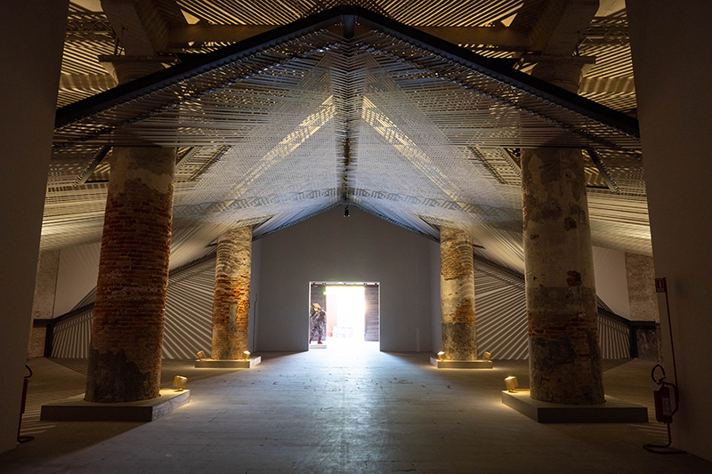 A gallery with six pillars and an artwork strung up using wide material straps.