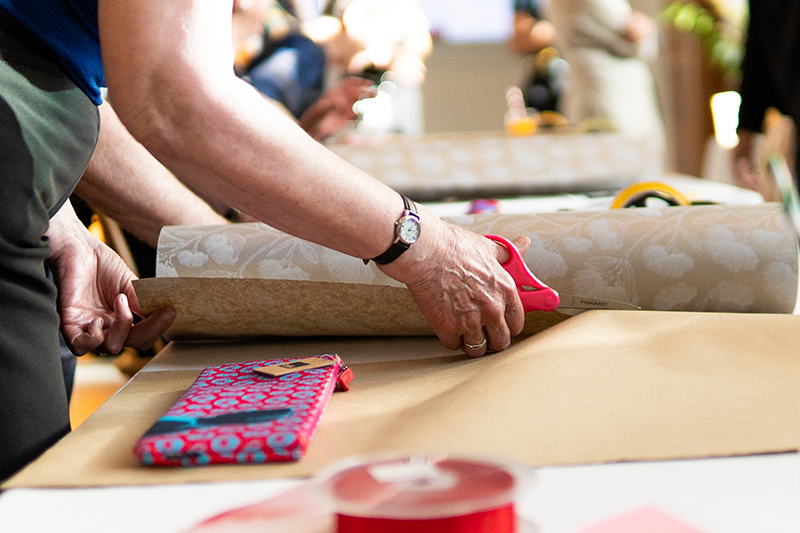 A close-up view of a person cutting wrapping paper with scissors. There is a wrapped gift in the foreground and some ribbon.