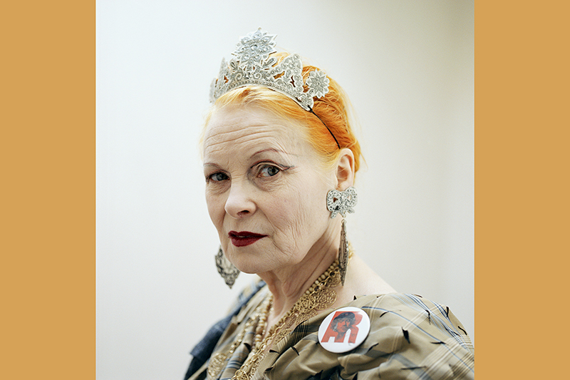 A head and shoulders photo of an older woman with dyed orange hair, a cardboard tiara, and large costume jewellery on. She is looking a the camera.