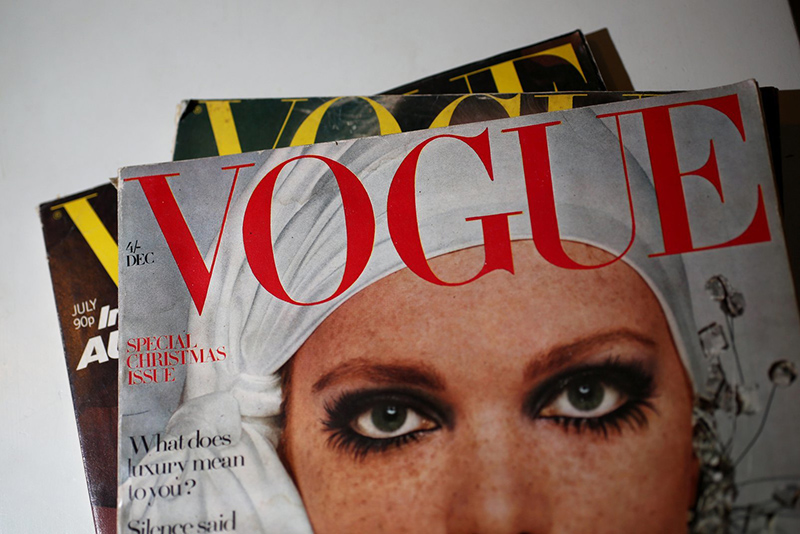 Several Vogue magazines in a pile with the one on top showing a close-up of a woman's face with heavily made up eyes.