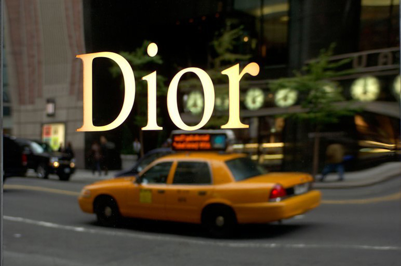 A window showing the reflection of a yellow cab and the word Dior printed in gold on the window.