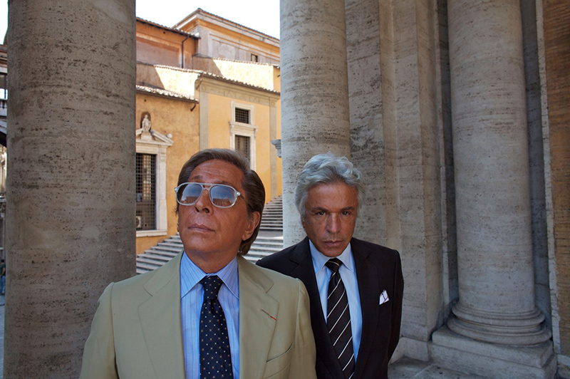 Head and shoulders of two men in suits walking between the pillars ourside a building in Rome.