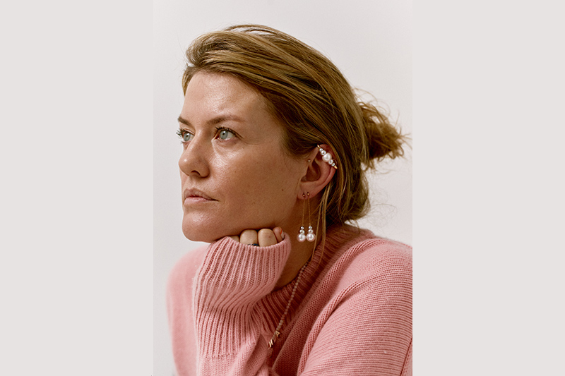 Head and shoulders photo of a woman with her hair tied up and wearing jewellery. She is looking off into the distance.