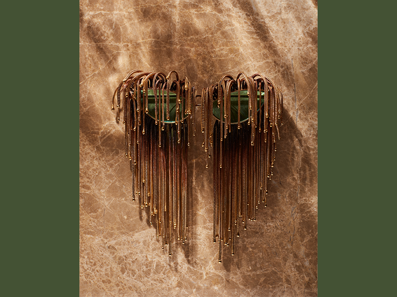 Gold rim cat-eye sunglasses with gold-bronze beaded fringe draping over the front and back of the green lenses. Sunglasses rest on a light brown, leather background.