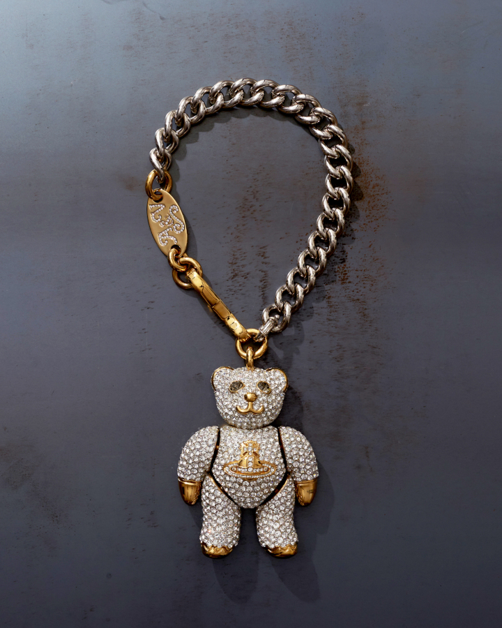 A diamond studded teddy hangs from a chunky silver chain necklace. It bears a golden Westwood symbol on its torso, with gold accents on the paws, nose and mouth. The gold clasp is engraved A&V.