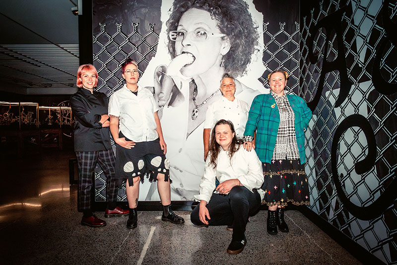 A diverse group of people wearing chefs jackets and punk fashion pose in front of a photographic mural of Vivienne Westwood eating a banana.