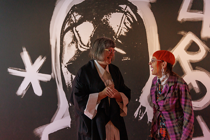 A photo of two people sharing a joke and laughing at each other. They are standing in front of a black wall with white designs painted on it.