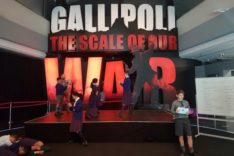 Several children in school uniform are milling about the front entrance to the exhibition Gallipoli The Scale of Our War at Te Papa.