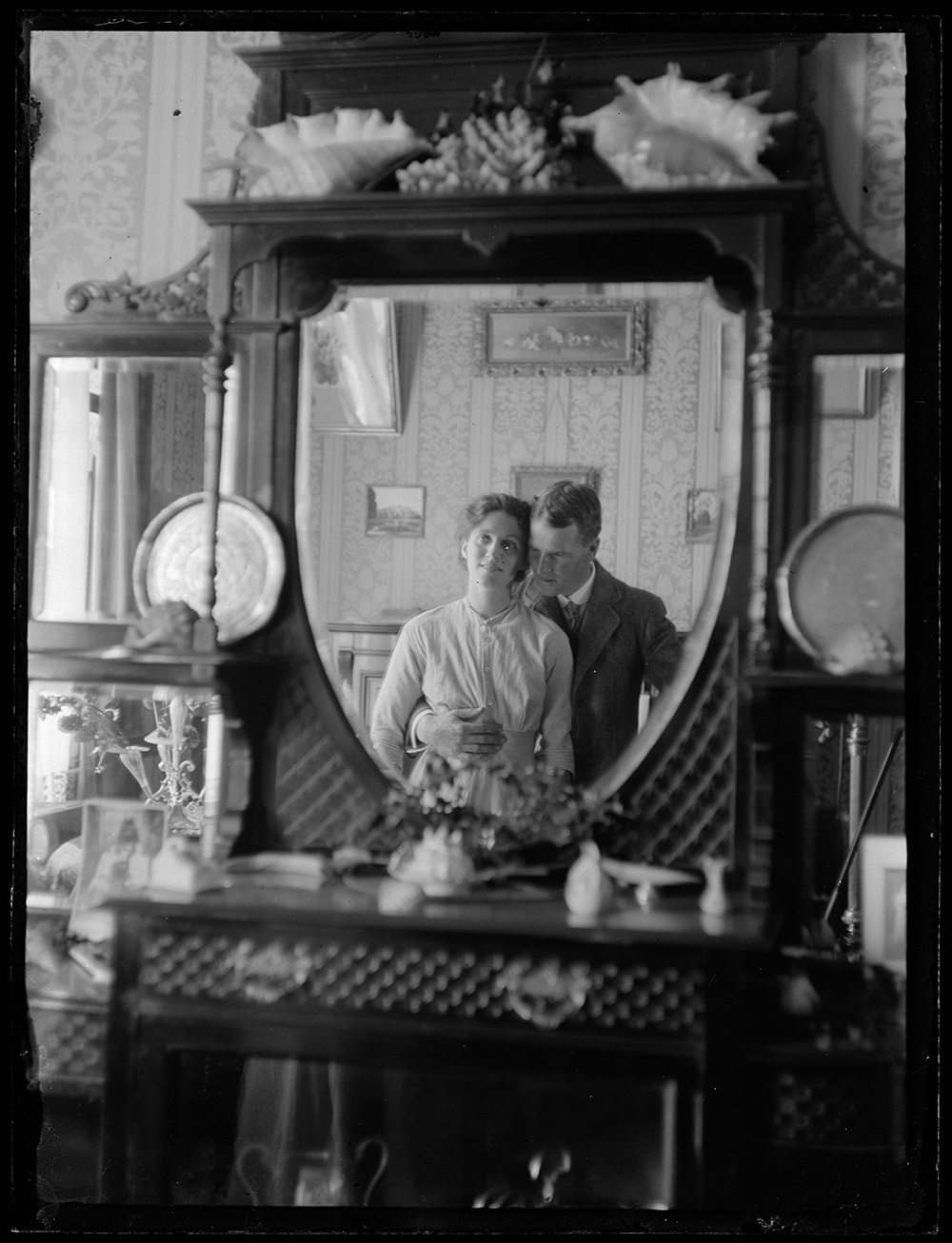 A photograph of a man and woman reflected in a dresser mirror. They are both facing the mirror and he has his arm around her waist.