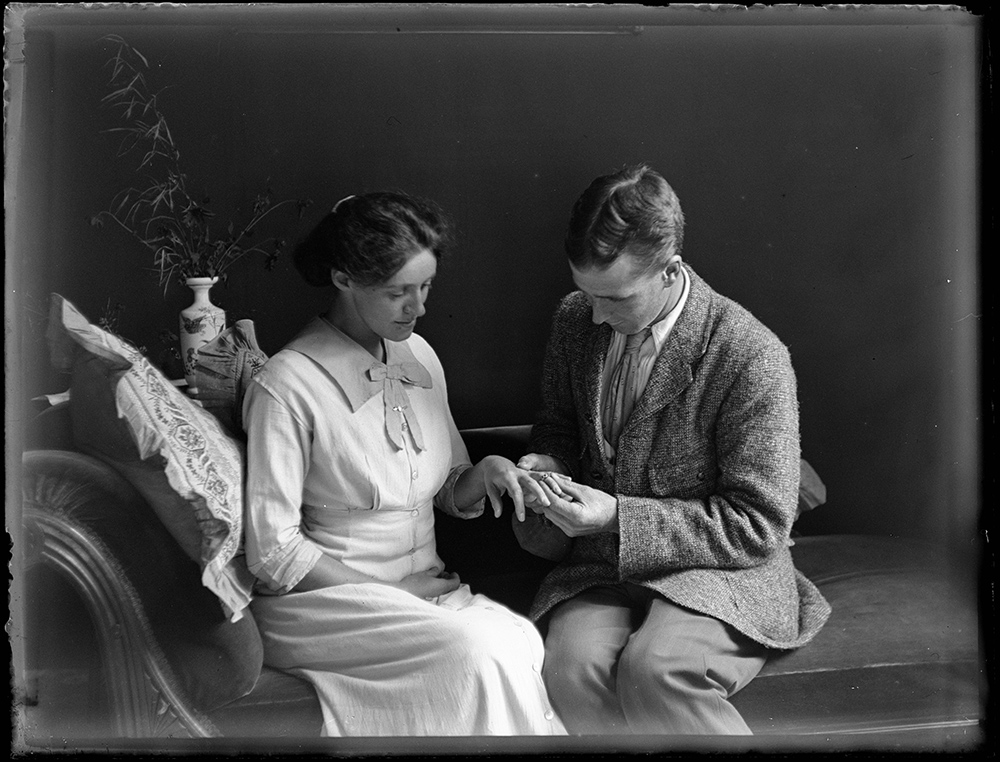 A black and white photograph of a man putting a ring on a woman's finger. They are sitting on a chaise longue.