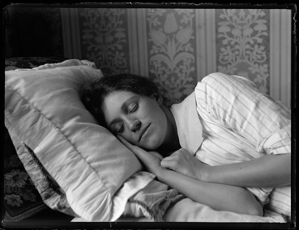 A black and white photograph of a woman sleeping on her side on a pillow. There is patterned wallpaper behind her.