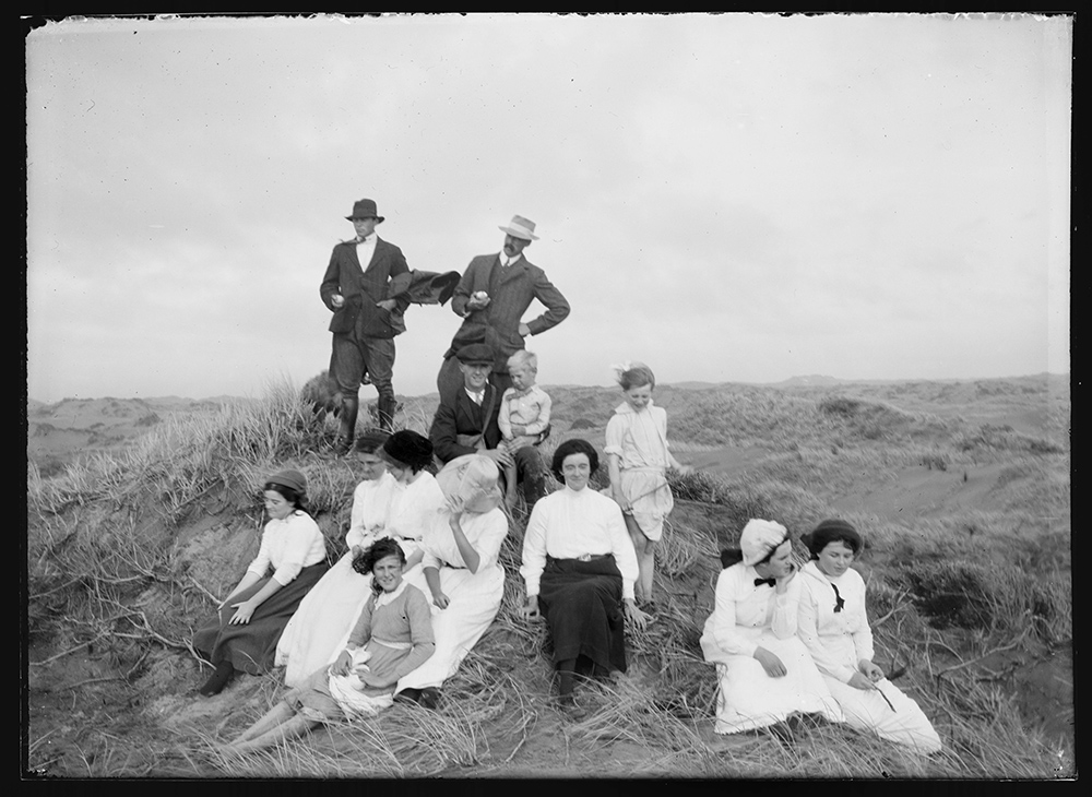 A black and white photo of several women and children sitting on a grassy sand dune. There are men in standing and sitting at the back.