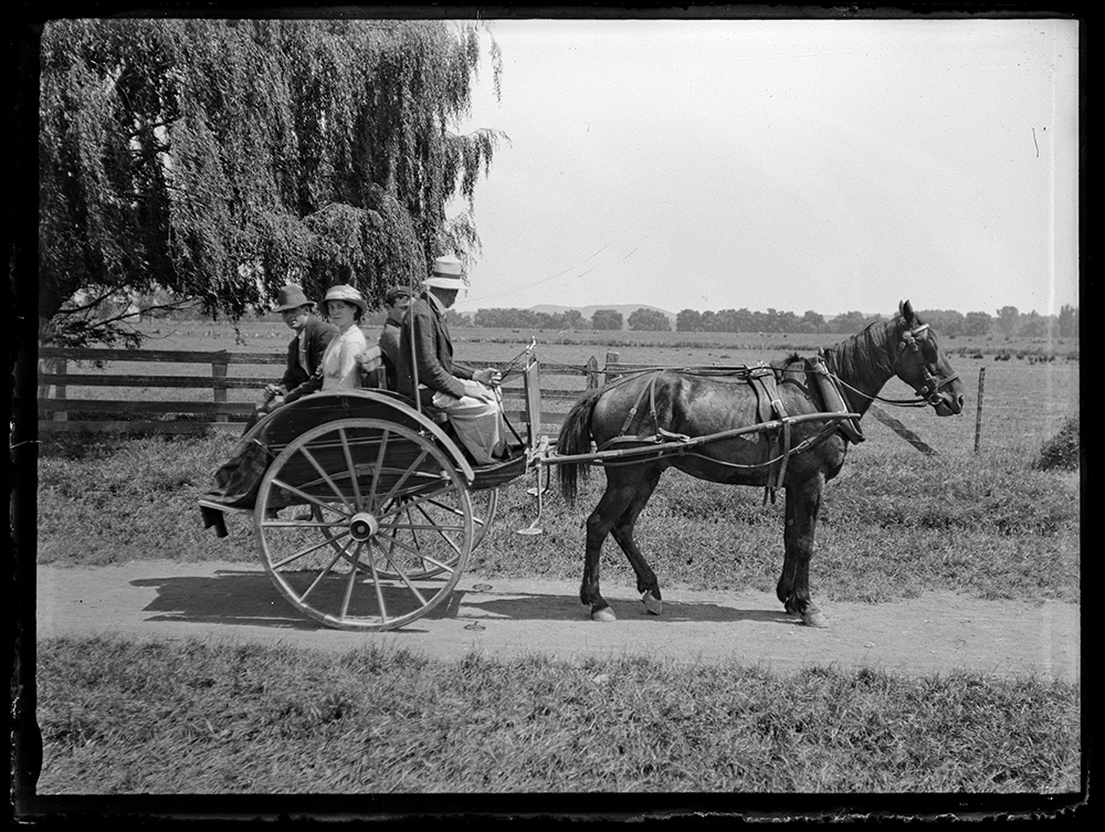 A black and white photo fo four people on a horse-drawn carriage. Three of the people are looking at the camera.