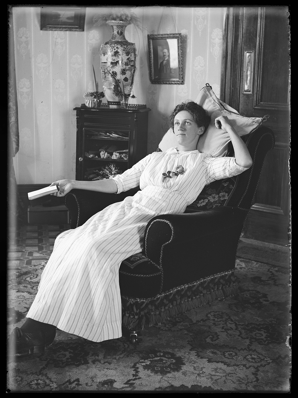 A black and white photograph of a woman in 1913 clothes reclining on a chair in a living room.