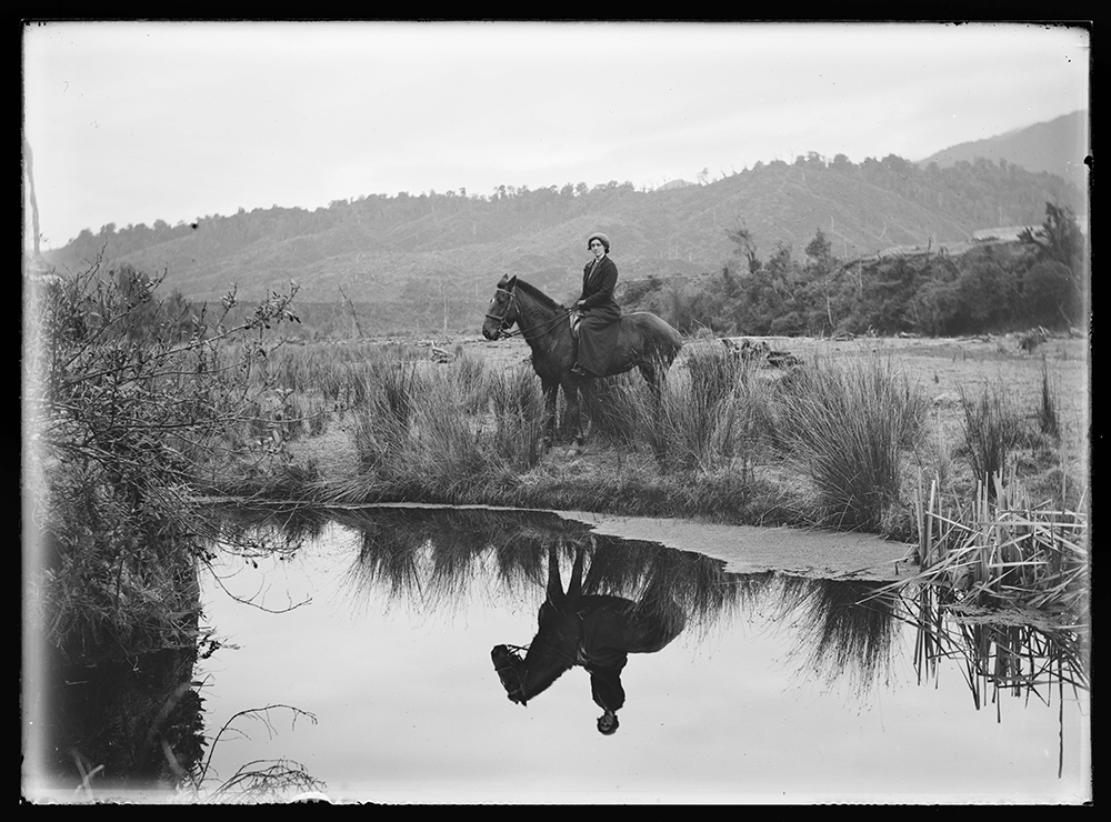 A black and white photo of a woman on a horse. They are both reflected in the calm pond in front of them.