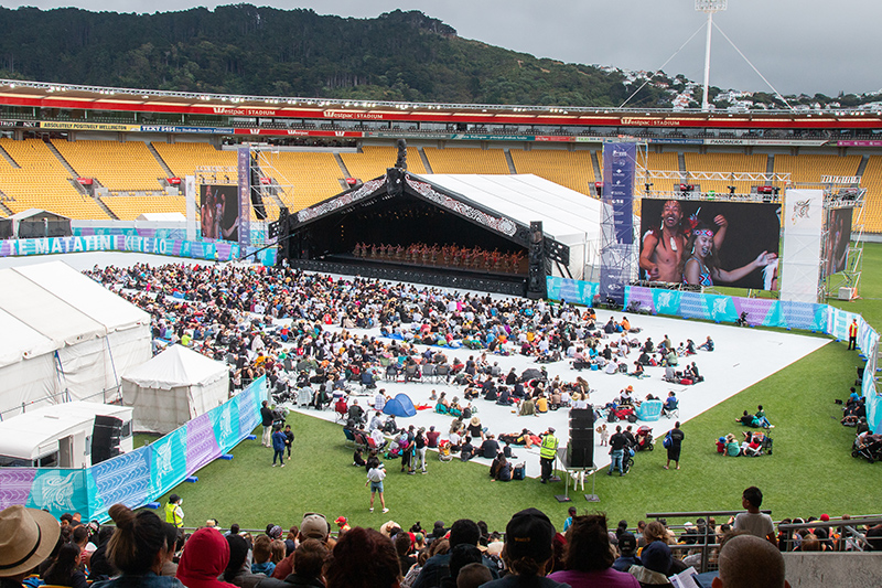 A view of a stadium with a crowd looking at a stage that is shaped like a Māori whare. There are large digital screens either side of the stage. The people are performing kapa haka, traditional Māori performance.