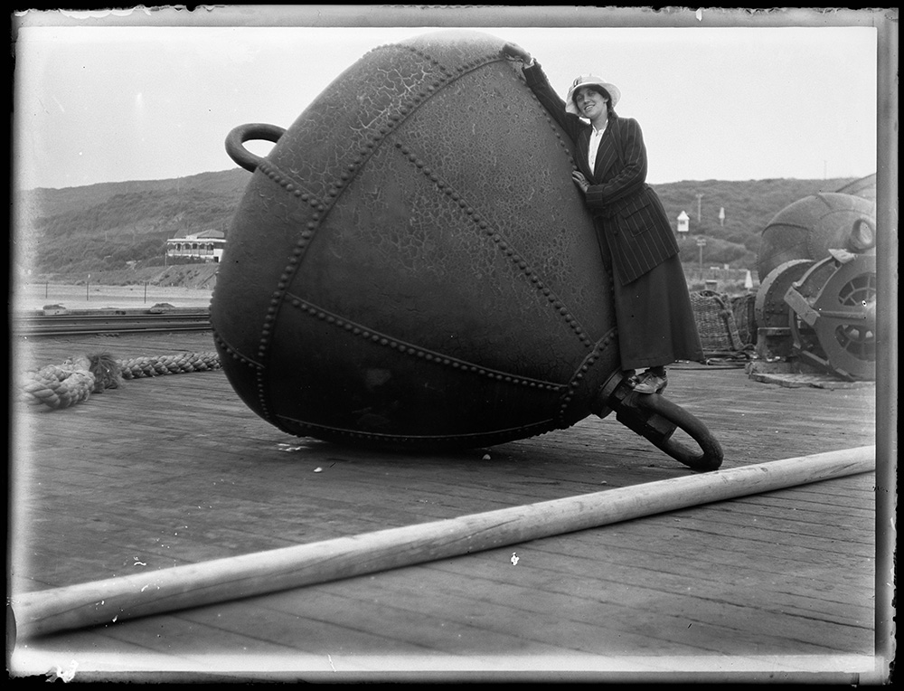 A black and white photograph of a woman standing on the side of a large ocean buoy on a pier.