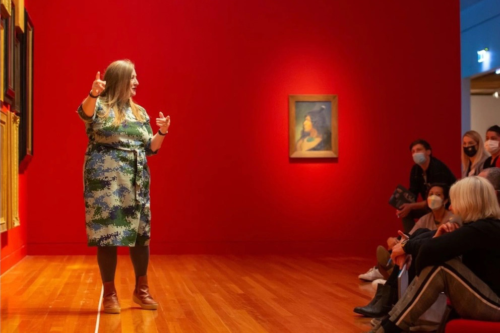 A woman in a dress is standing in front of some art and pointing to it as she talks to some people who are seated in front of her.