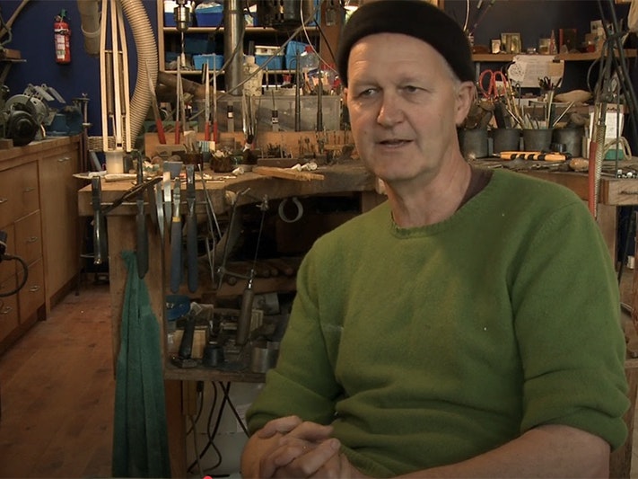 Still from Interview with Warwick Freeman. Te Papa A man in a woollen cap and green jersey is talking to someone just off camera. In the background is his jewellery studio.
