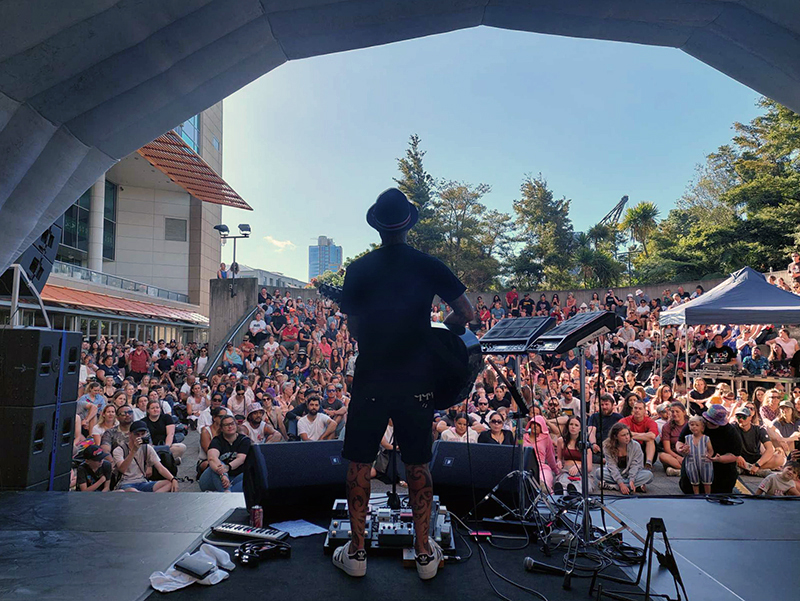 A man is standing with his back to the camera and is playing a guitar to an outside audience. There is an inflatable stage cover around him.