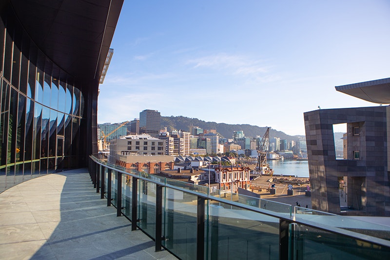 A photo from a balcony on a modern building. The view is part of the city of Wellington showing a part of Te Papa and the harbour as well as Te Ahumairang hill.