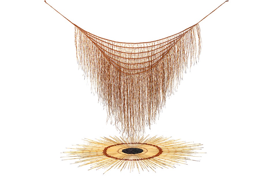 An art work with a woven harakeke (flax) cape is hanging over another woven artwork arranged in a circle on the white ground.