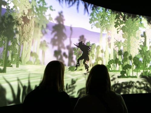 Māui explores Aotearoa at the entrance to Te Taiao, 2019. Te Papa Two silhouetted people are watching a screen where another silhouetted person is creeping through the forest.