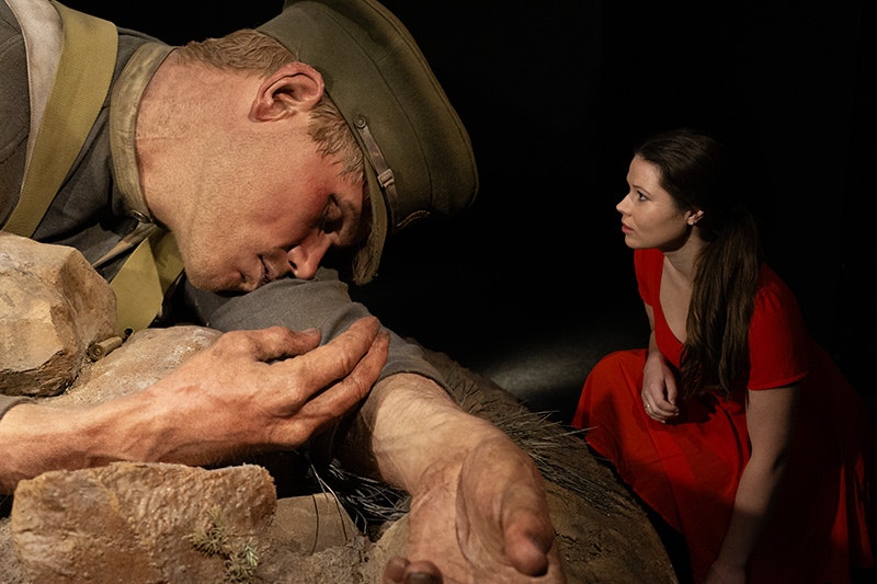 A woman in a red dress is crouched next to a giant model of a soldier.