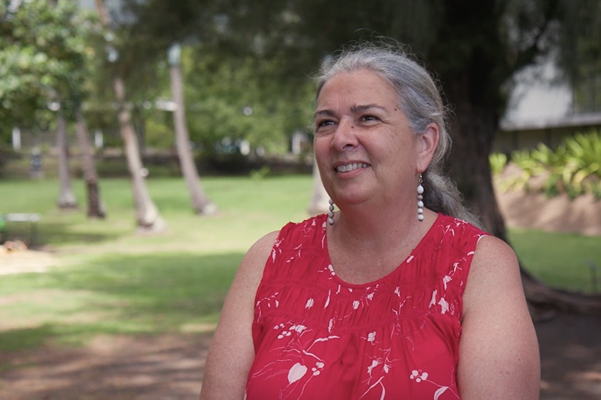 A woman with her grey hair tied back in a ponytail and wearing a sleeveless red top is standing on some grass smiling at someone off camera.