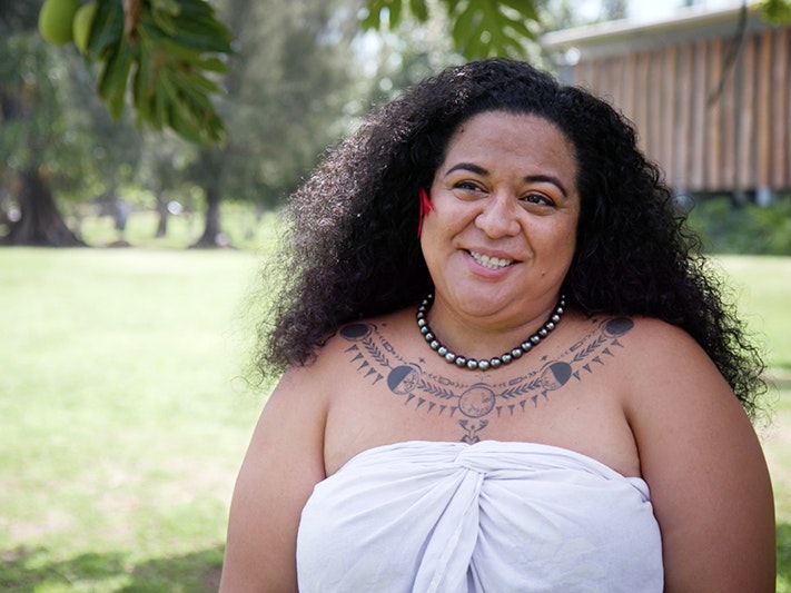 Still from the interview with Hinatea. Te Papa A woman with long curly black hair and wearing a wrap dress. She has a tattoo around her neckline.