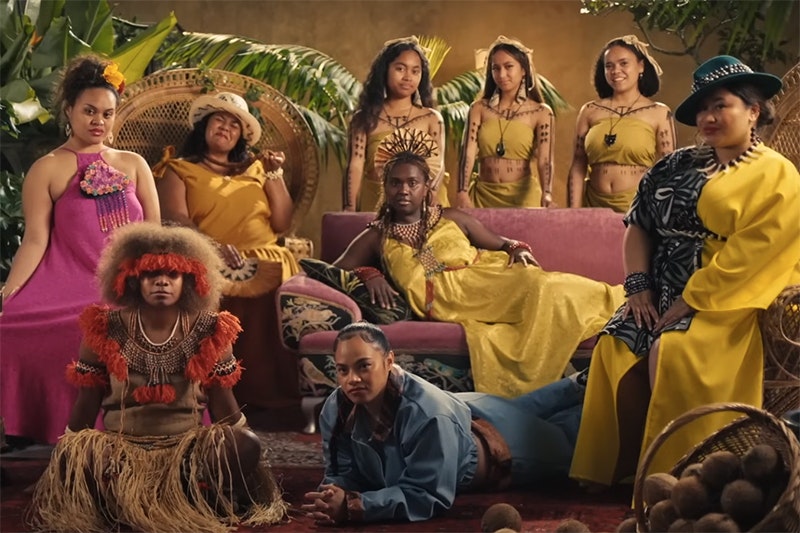 Nine women wearing pasifika clothing are standing or seated around or on a sofa looking at the camera.