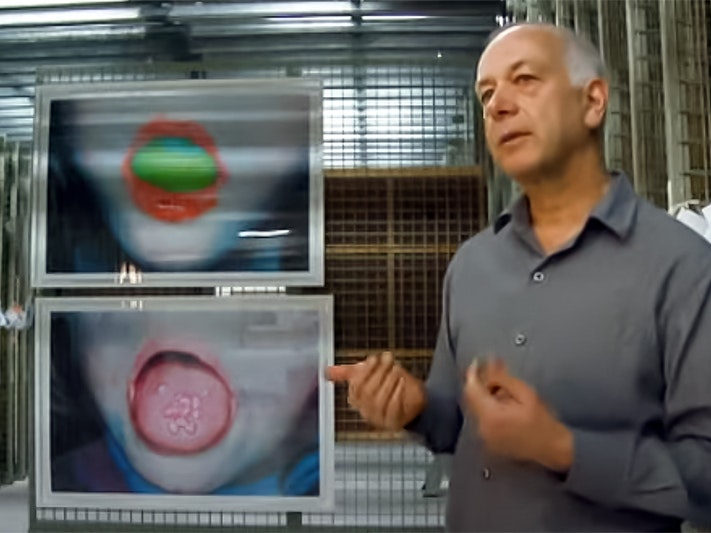 Still from Tales of Te Papa - Ruby's Room. A man in a grey shirt is standing in front of two large photographs of children's open mouths. One of the mouths has something bright green in it. The oher is sticking out it's tongue and has something pink on it.