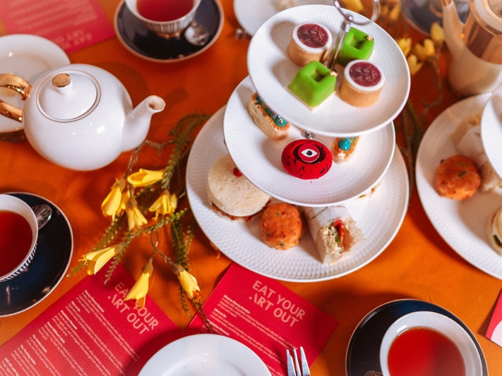 Eat Your Art Out, 2025. Photo by Victoria Saunders An image of an orange-red table with tea and teapots along with three-tierd plates with small cakes.