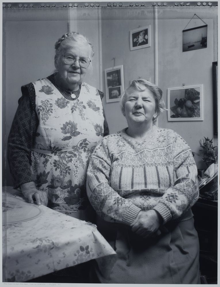 A black and white photo of two older woman smiling at the camera. One is standing by another sitting down.