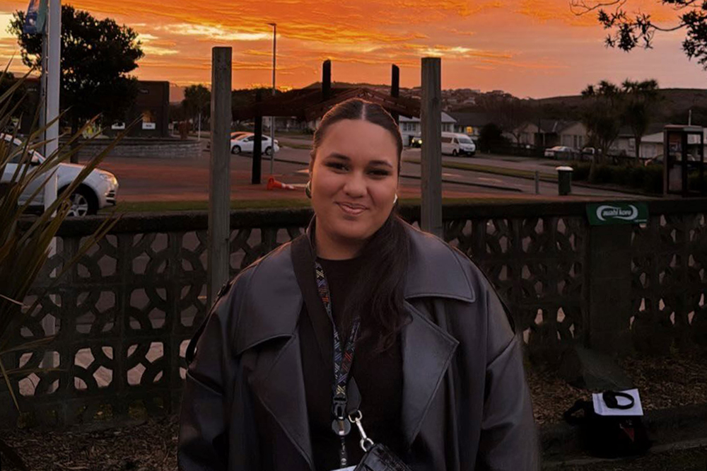 A woman in black clothing and a black jacket is standing outside in front of a carpark at sunrise.