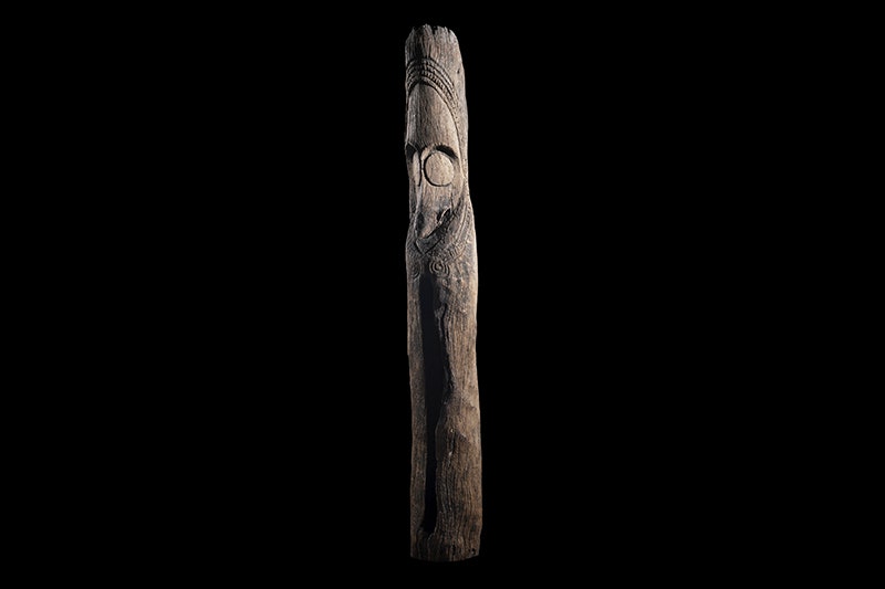 A tall carved wooden pole with a face in the top half and a vertical slit in the hollow bottom half.