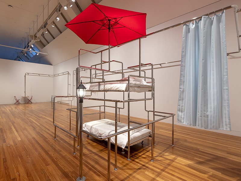 Yona Lee, Kit--set In-transit, 2020. Installation view (detail), 2025. Purchased 2021. Te Papa (2021-0030-1) A large art gallery space with an artwork made from chrome bars, a red umbrella, and some beds set into the bars.