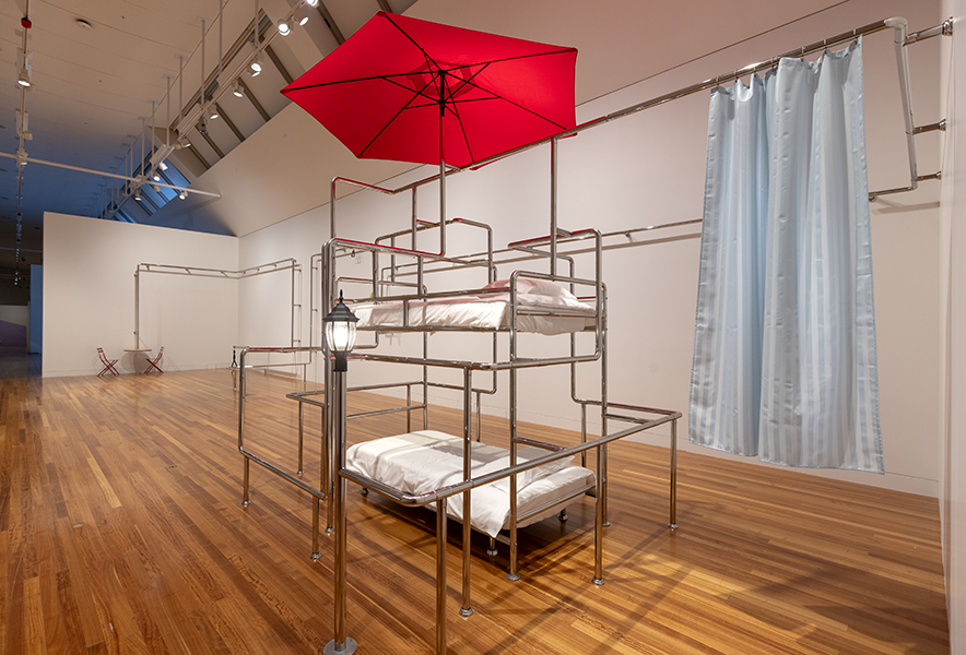 A large art gallery space with an artwork made from chrome bars, a red umbrella, and some beds set into the bars.