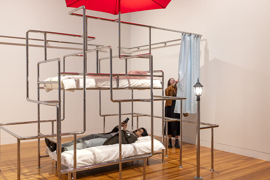 A person is lying down on a bed in a gallery that is surrounded by chrome piping. There is another person pulling a shower curtain standing next to them.